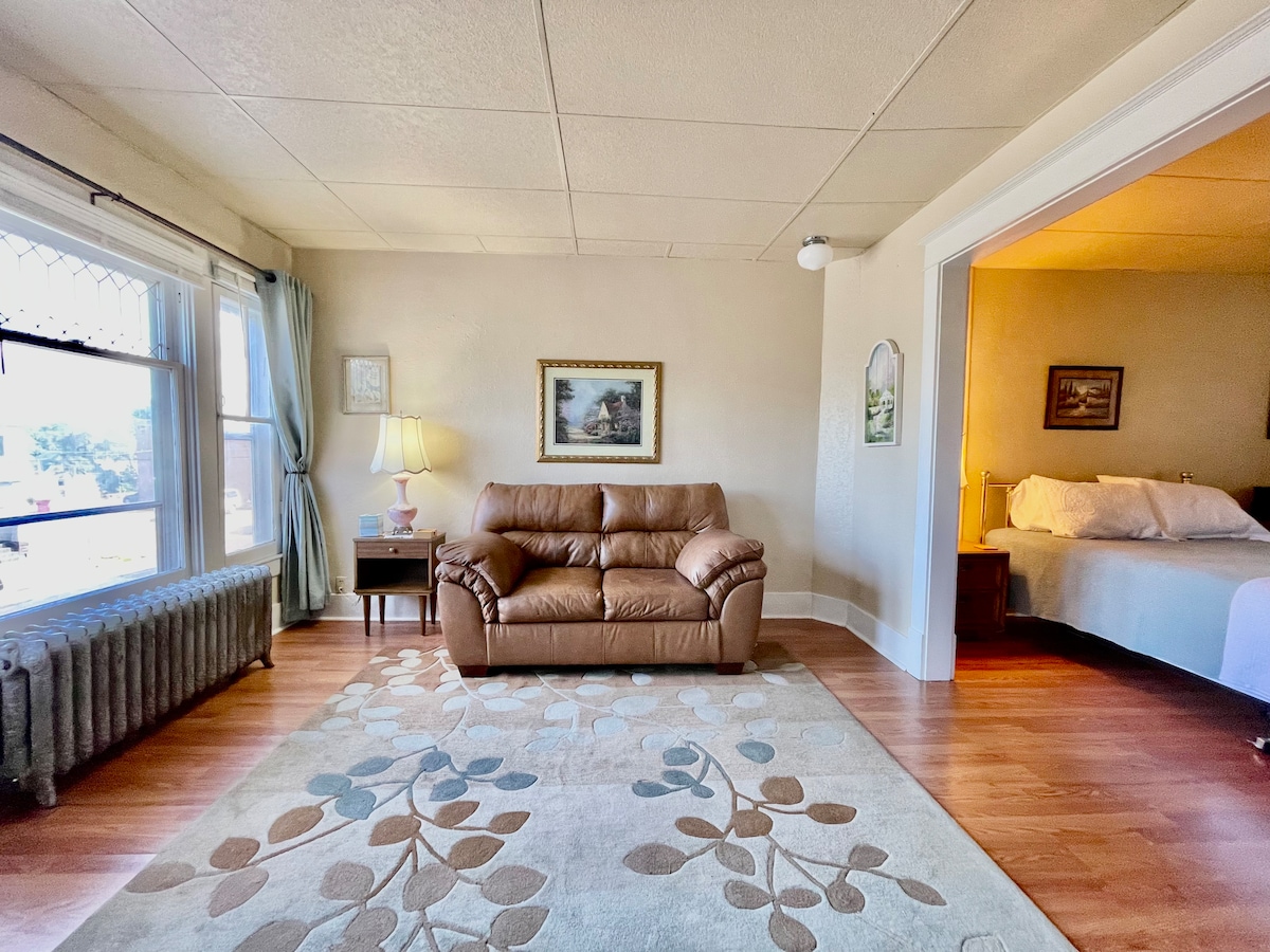 A living area features a comfortable loveseat facing a window, with natural light illuminating the space. A decorative rug with a floral pattern covers the hardwood floor. A small lamp and framed artwork are visible, providing a sense of homely decor.