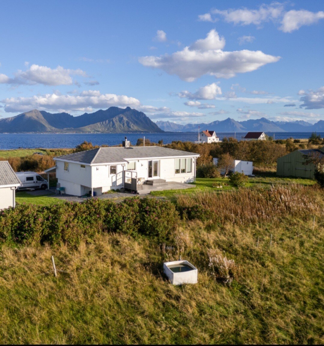 The property is situated on a peninsula surrounded by lush greenery and features a well-maintained exterior. Scenic views of mountains and the sea are visible in the background, with a clear sky dotted with clouds. The area is tranquil and natural.