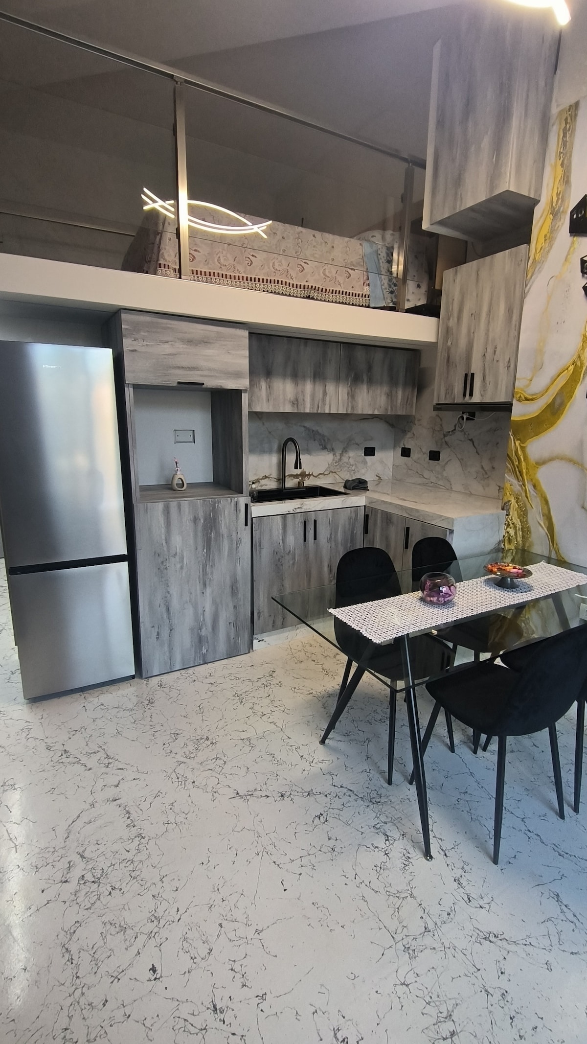 A modern kitchen area features sleek cabinetry with a marble backsplash. A stainless steel refrigerator stands alongside a compact cooking space. A glass dining table with four black chairs is positioned nearby, enhancing the efficient layout of the studio apartment.