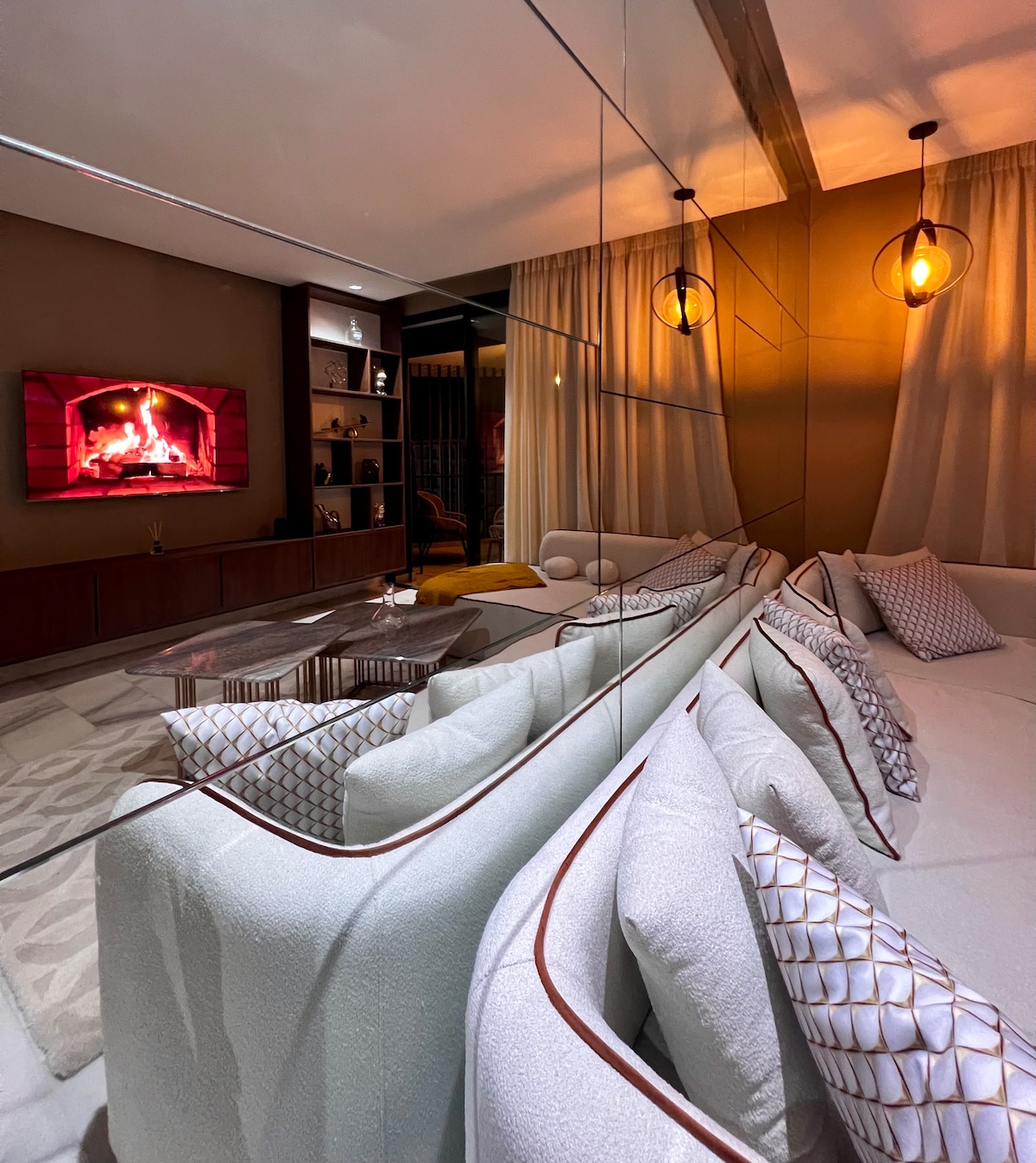 A modern living area features a comfortable sectional sofa adorned with patterned cushions. A wall-mounted television displays a glowing fireplace, adding warmth to the space. Soft lighting is provided by pendant lamps, while sheer curtains frame a window, enhancing a cozy atmosphere.
