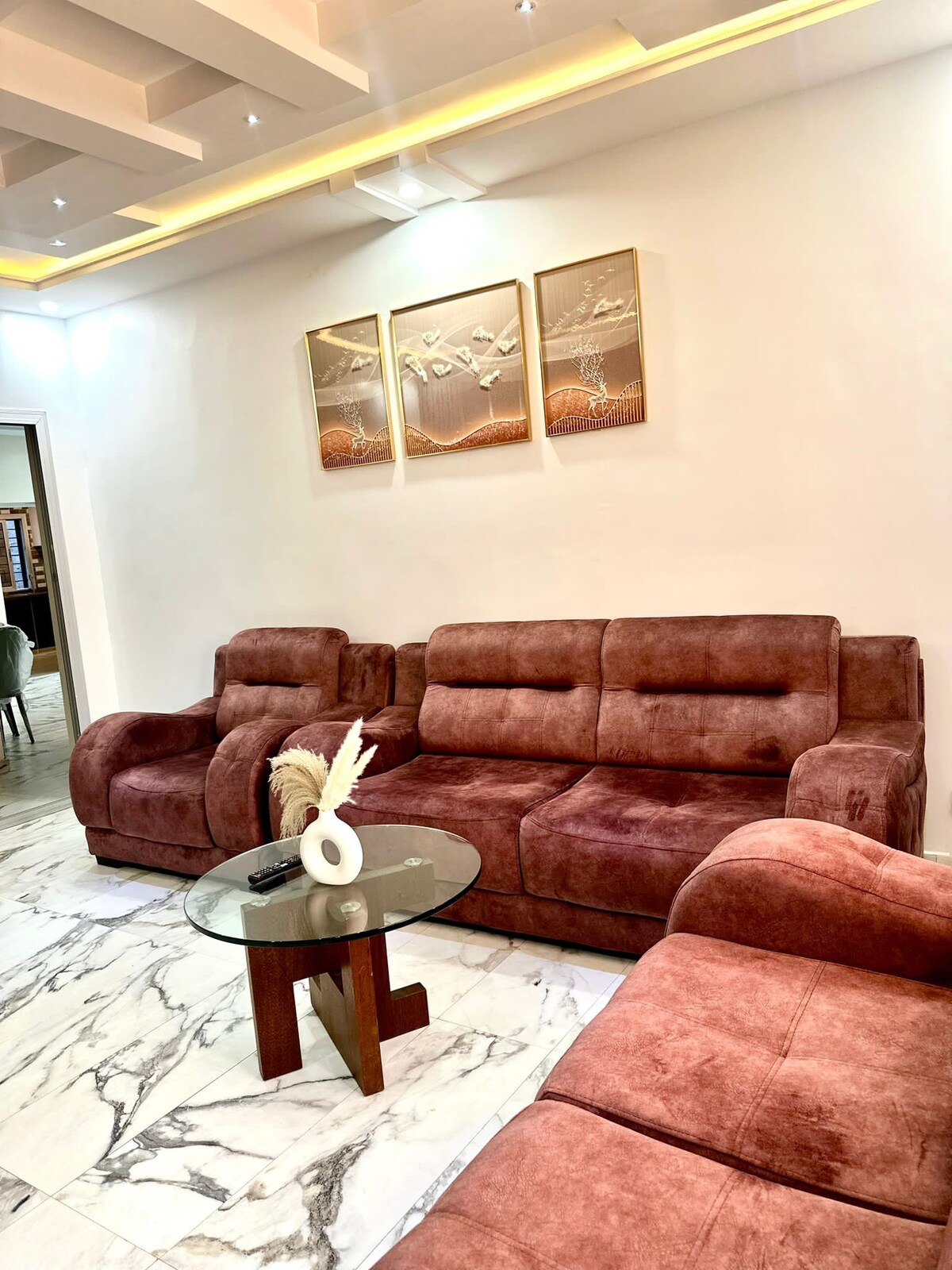 A spacious living area features a plush, brown sectional sofa arranged around a low glass table. Above the sofa, three decorative art pieces are mounted on the white wall, while soft lighting enhances the modern atmosphere. The marble flooring adds a touch of elegance.