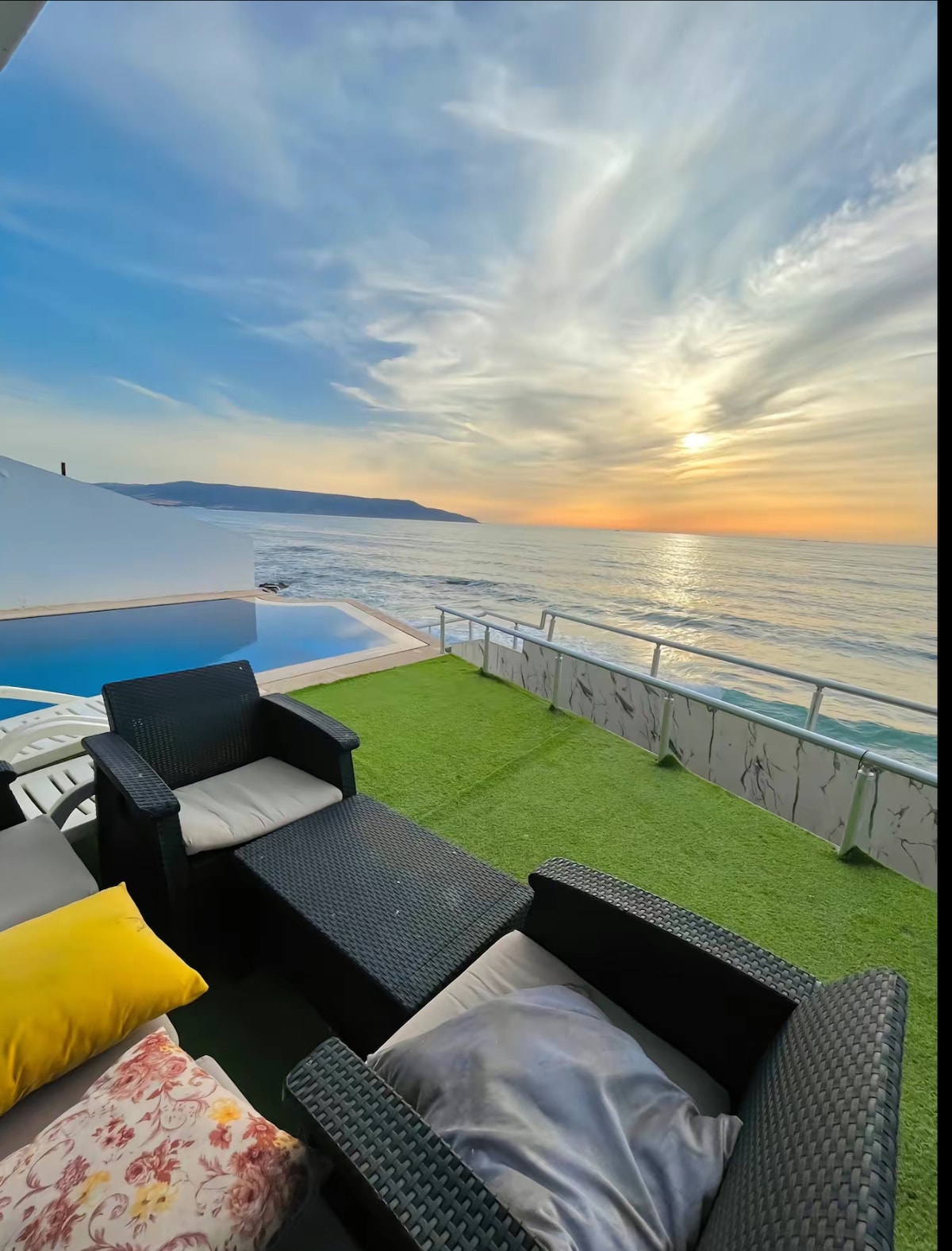 An outdoor seating area is equipped with dark wicker chairs and a small table, set against a backdrop of the ocean and a sunset sky filled with soft, colorful clouds. A private infinity pool is visible, with a grassy area enhancing the tranquil atmosphere.