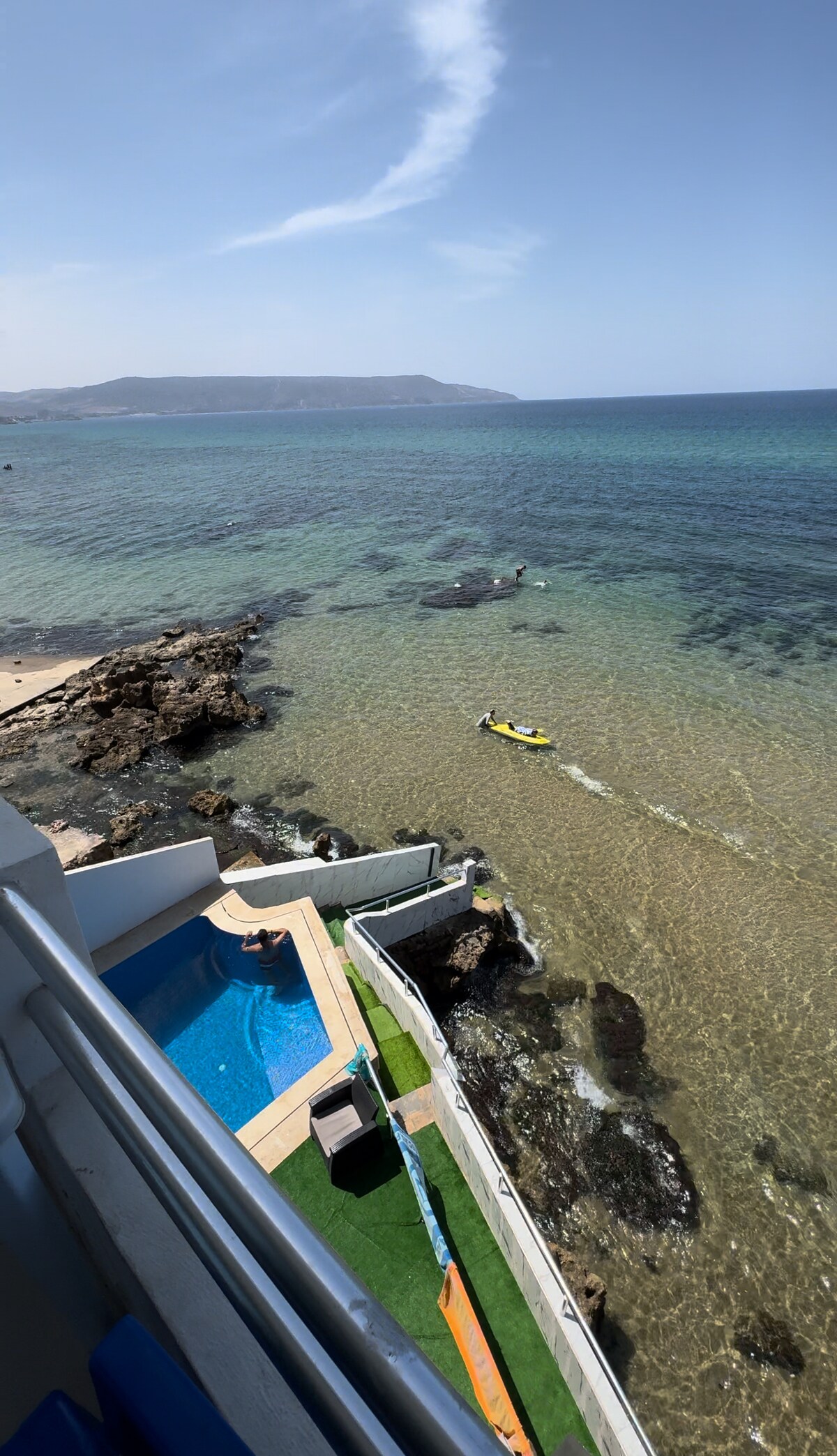 A stunning view of the sea is captured from a balcony, featuring clear waters and rocky shoreline. The infinity pool is visible, along with a sunbathing area on lush green grass. A small watercraft is seen on the water, emphasizing the coastal ambiance.