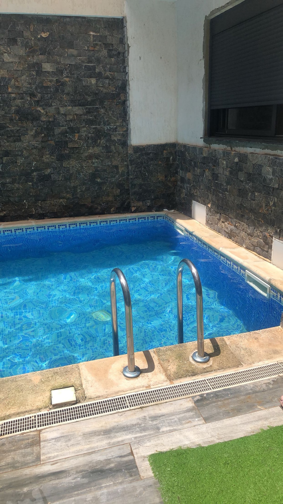 A private swimming pool is framed by textured stone walls, showcasing a vibrant blue water surface. Two stainless steel ladders provide access to the pool, which is bordered by a tiled edge. Natural light enhances the inviting atmosphere.