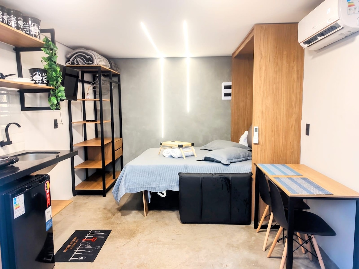 A compact studio features a spacious bed with neutral linens and bedside tables. A dining area with a small table is positioned near the kitchen, which includes a fridge and open shelving. The decor is complemented by ambient lighting and air conditioning unit on the wall.