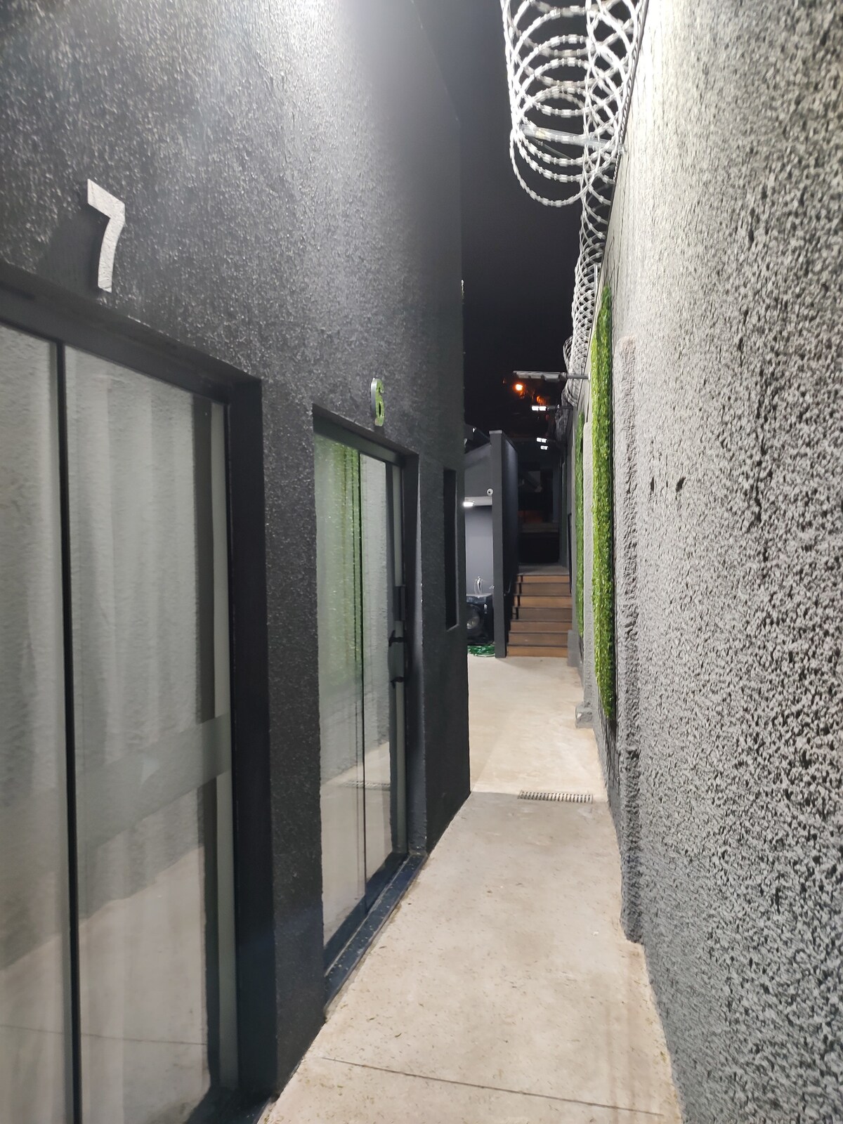 A narrow corridor is flanked by black walls and features glass doors at the sides. Soft lighting illuminates the space, enhancing visibility. Barbed wire is seen along the top of the walls, and a textured surface adds to the modern aesthetic.
