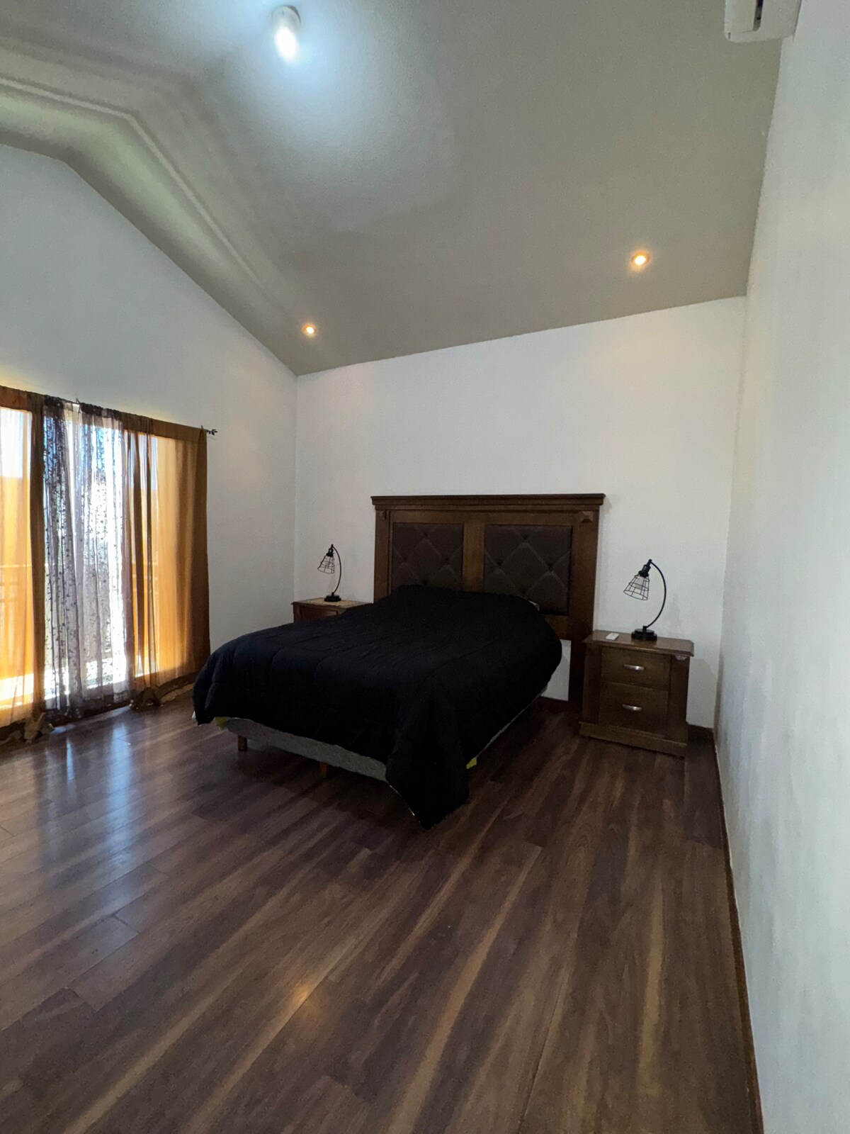 A well-appointed bedroom is presented with a spacious bed dressed in dark bedding. Wooden nightstands are located on either side, each featuring a table lamp. Natural light enters through floor-to-ceiling curtains, contrasting against the wooden floor and white walls.