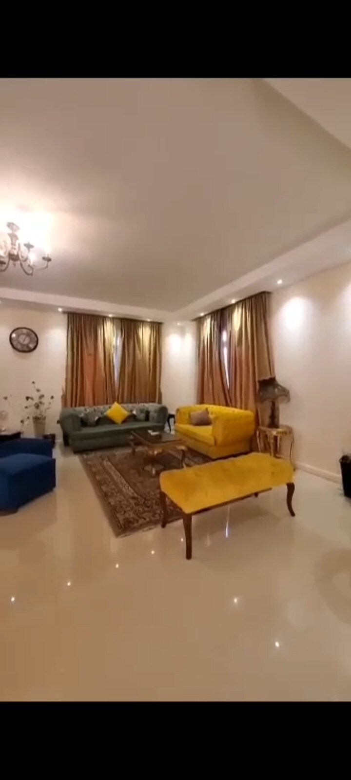 A spacious living area features a combination of blue, green, and yellow seating, complemented by a decorative area rug. Natural light filters through long, golden curtains, while a chandelier adds subtle illumination. Decorative accents and a wall clock enhance the cozy ambiance.