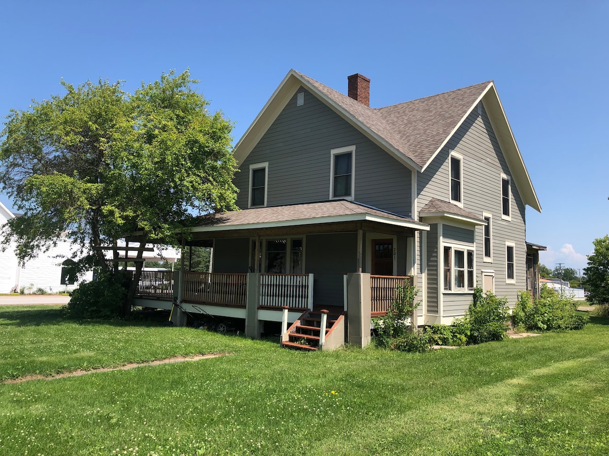 A charming two-story house is bordered by a green lawn and mature trees. The front porch features a wooden railing and steps leading to manicured gardens, creating a welcoming entrance. Sunlight illuminates the exterior, highlighting its restored siding and architectural details.
