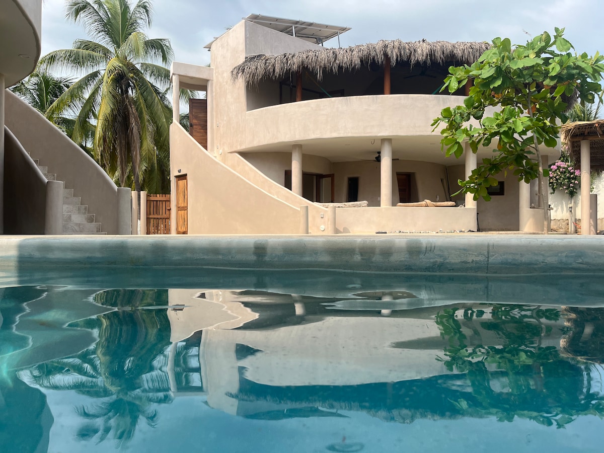 A serene outdoor space features a clear blue pool reflecting the architectural design of the villa. The building showcases a thatched roof above spacious balconies and large windows framed by palm trees, providing an inviting connection to the lush surroundings.