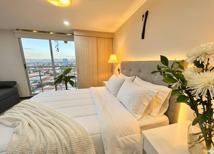 Dormitorio acogedor y luminoso con cama confortable, ropa de cama impecable, detalles decorativos y una hermosa vista panorámica de Bogotá.