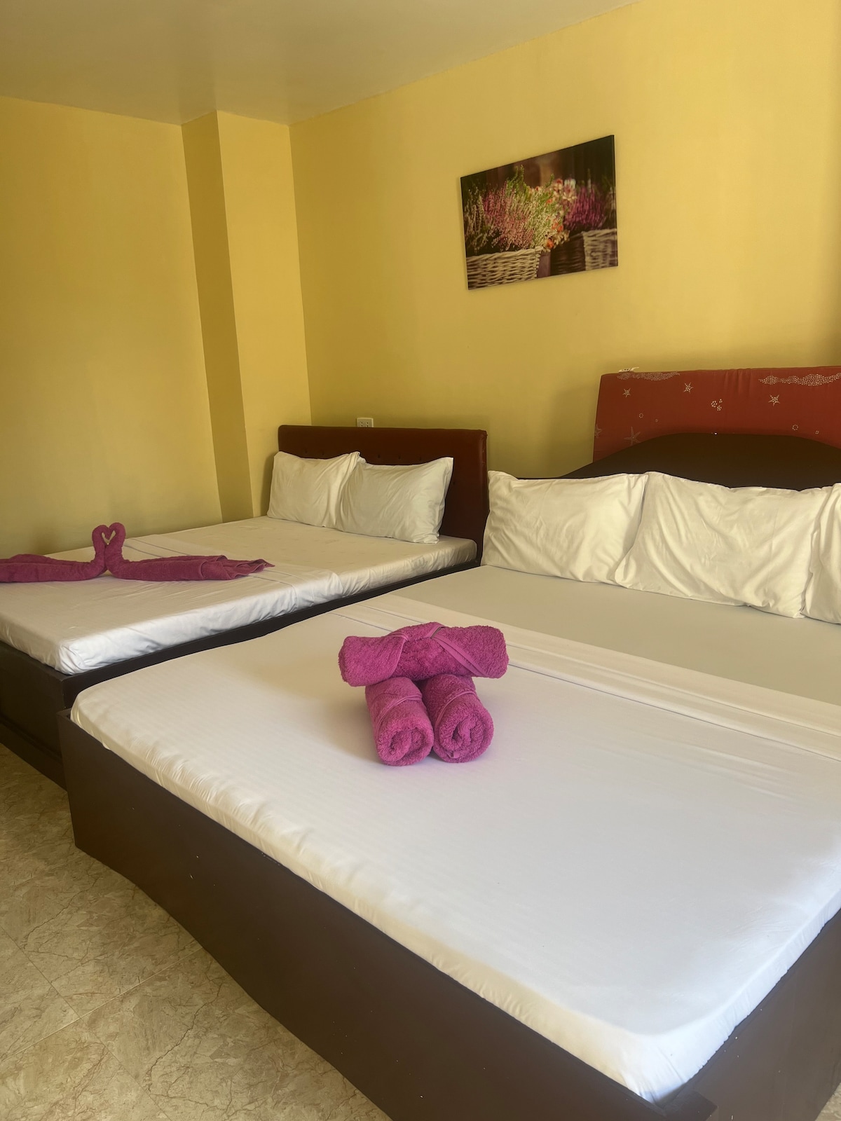 Two spacious beds are arranged in a light-filled room with soft yellow walls. Each bed is adorned with white linens and complemented by colorful towels. A framed floral artwork is displayed above the headboard, adding a subtle decorative touch.