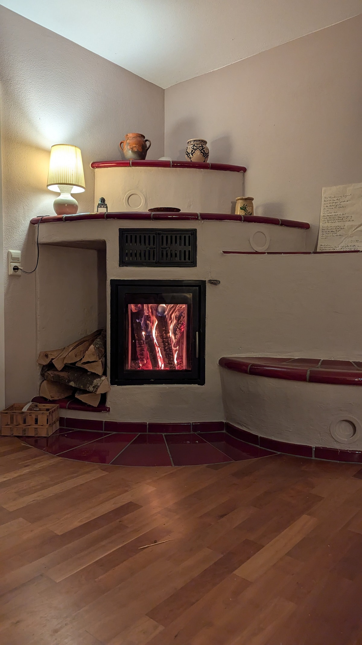 A cozy corner features a wood-burning stove with visible flames, surrounded by a rustic tile hearth. Shelves with decorative items and a warm lamp add subtle illumination. A wooden basket holds logs, and wooden flooring enhances the inviting atmosphere.