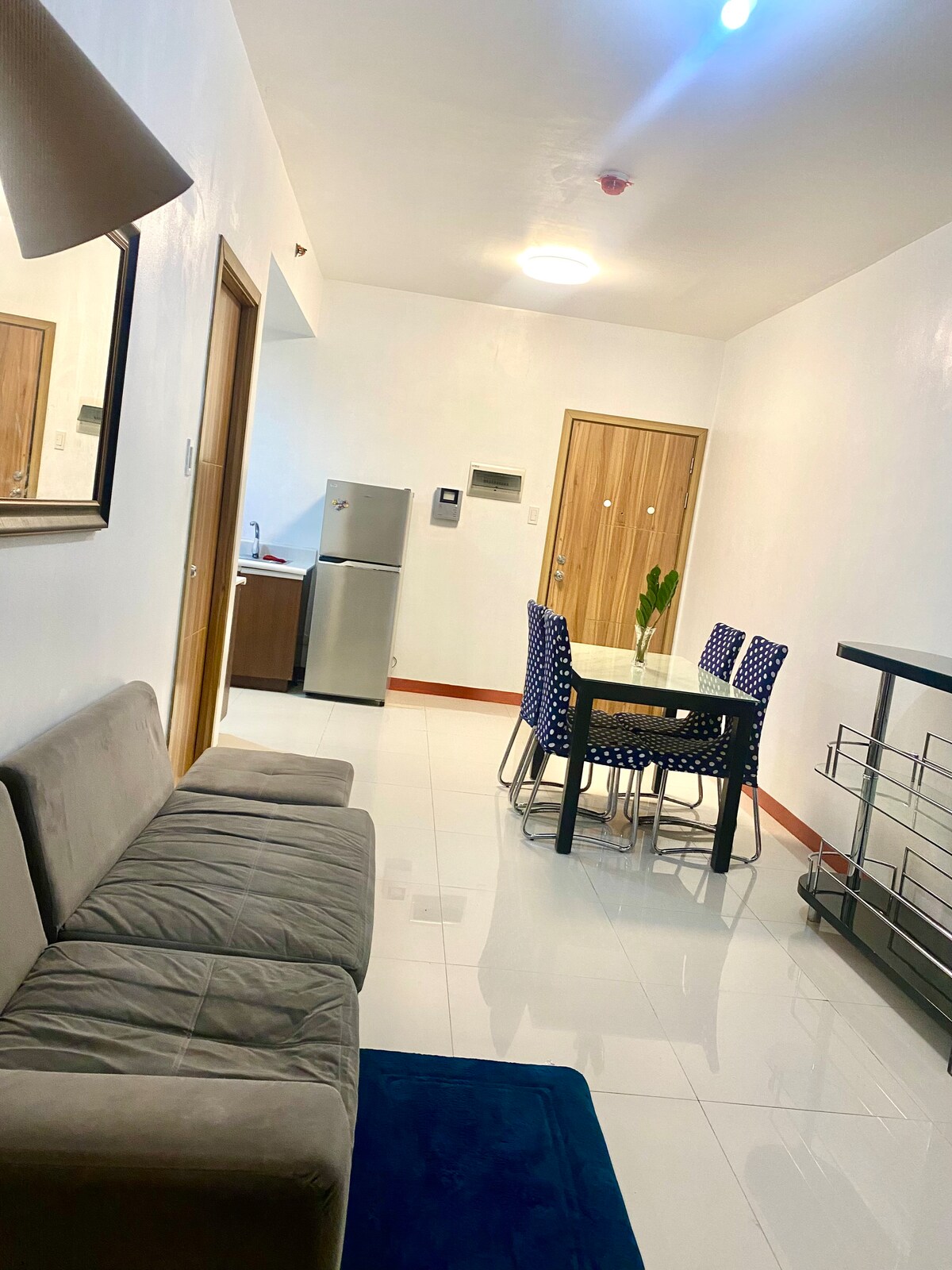 A functional living area features a comfortable sofa and a dining table surrounded by six chairs. A mirror reflects the room's bright lighting. The kitchenette is seen in the background, equipped with modern appliances. Neutral-colored walls and tiled flooring create a spacious feel.