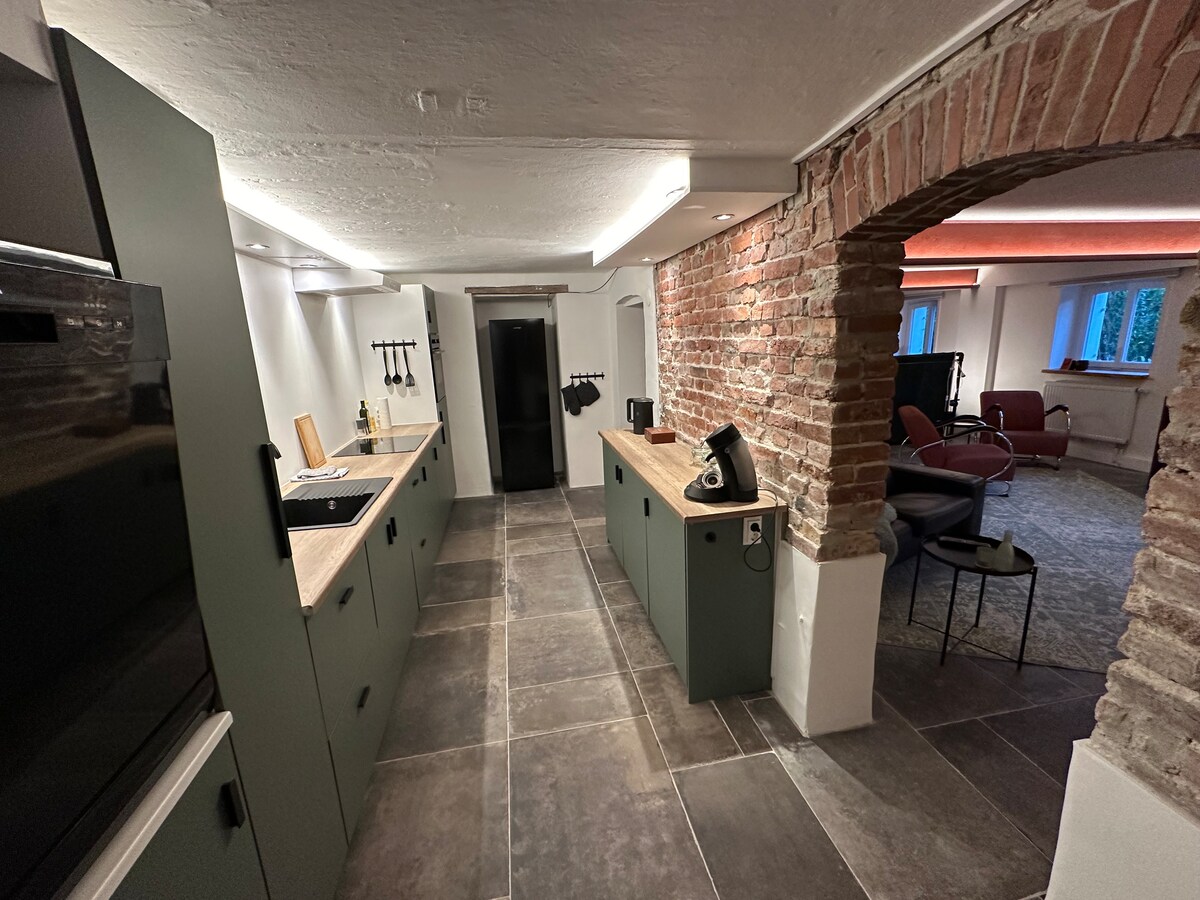 A spacious kitchen area is presented with modern cabinetry and a long countertop made of natural wood. Exposed brick walls add character, while stainless steel appliances, including a refrigerator, are integrated into the design. Soft lighting enhances the inviting atmosphere of the space.