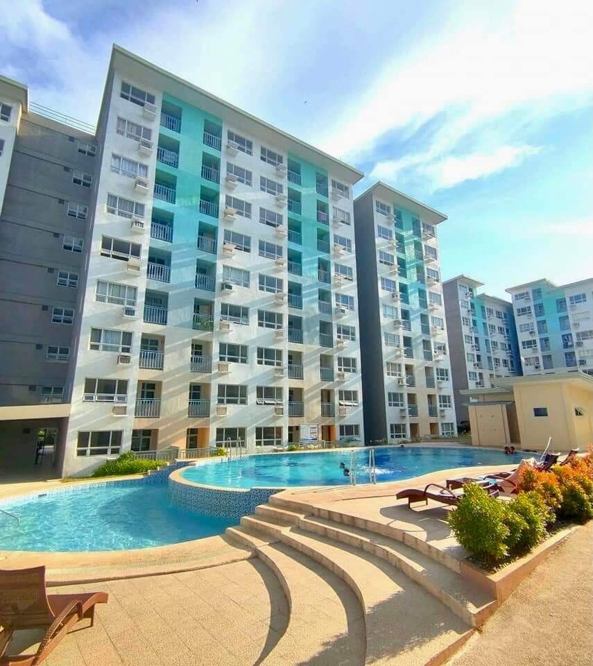 A modern multi-story building features a series of balconies overlooking a tranquil swimming pool. Stone steps lead down to the inviting pool area, surrounded by lounge chairs and greenery, set against a backdrop of clear blue skies.