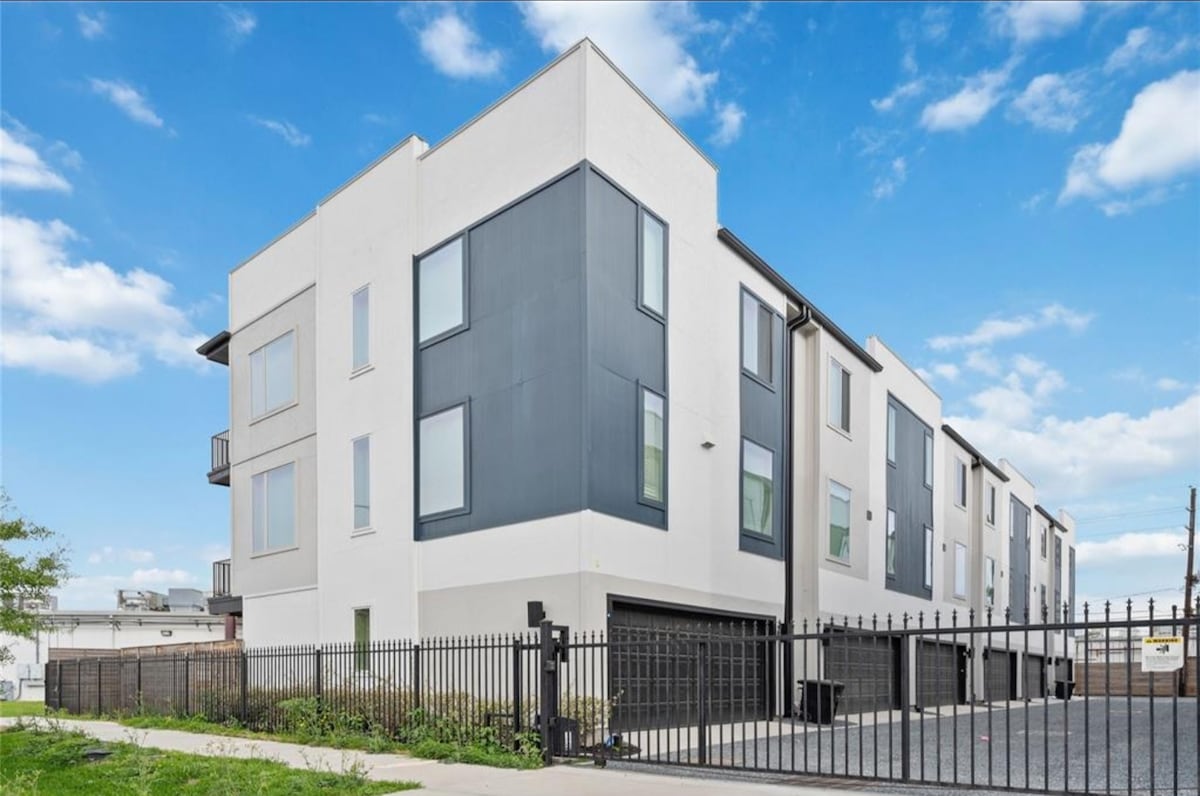A modern three-story townhome is showcased, featuring a mix of light and dark exterior panels. Large windows are present on each floor, allowing for ample natural light. A gated entrance provides security, and a spacious driveway leads to the garage below.