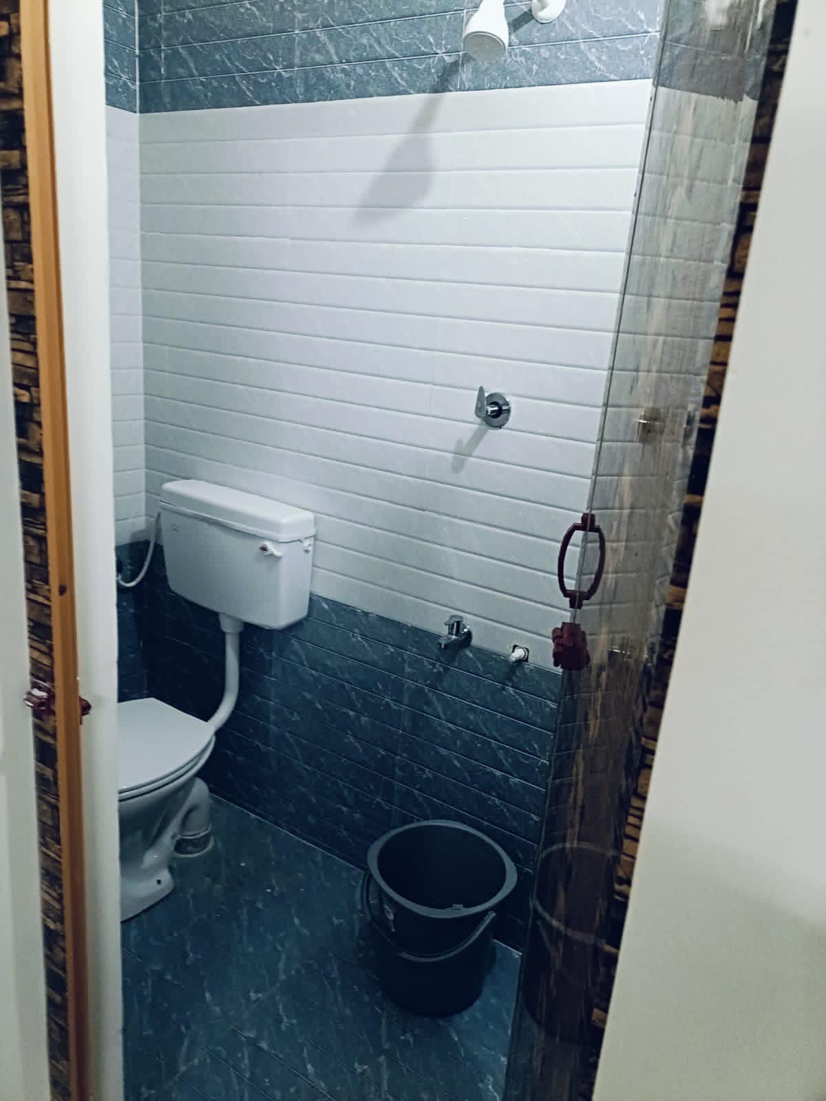 A clean and functional bathroom is presented, featuring a modern toilet positioned against white tiled walls. Shower fixtures are installed above, and a bucket is positioned on the floor. The blue tiled flooring complements the overall design.
