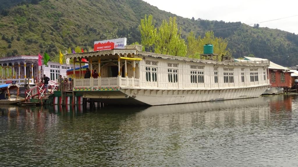 A boat house is anchored on calm waters, displaying intricate wooden carvings along its exterior. Lush green hills rise in the background, contrasting with the vibrant colors of nearby structures. The scene conveys a sense of tranquility, inviting exploration of the natural surroundings.
