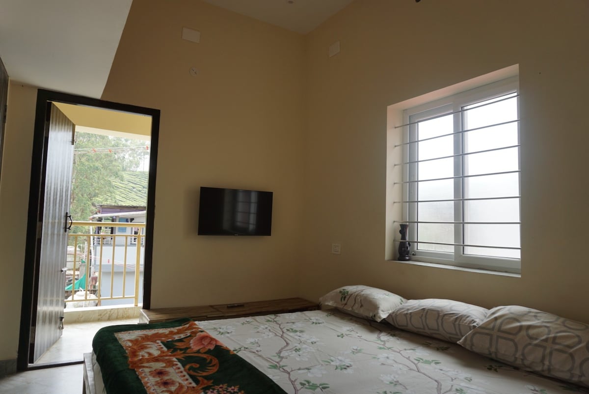 A bright room features a spacious bed with decorative bedding, accompanied by several pillows. A flat-screen television is mounted on the wall opposite the bed. Natural light enters through a window, and a balcony view is visible through an open door, enhancing the room's openness.