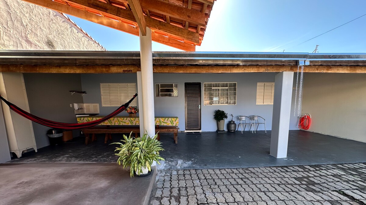 An outdoor area is shown, featuring a covered patio with a hammock and wooden seating. There are plants positioned on both sides, with a small table and chairs for relaxation. Light enters through several windows along the facade, enhancing the inviting space.