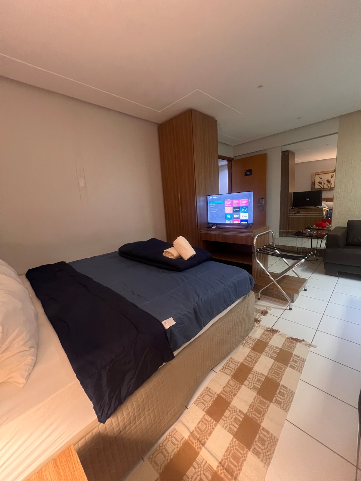 A well-appointed room features a spacious bed dressed in dark bedding, with crisp white pillows. A flat-screen TV is mounted on a wooden panel, and a small dining table with clear chairs is positioned nearby. Soft lighting creates a calm ambiance.