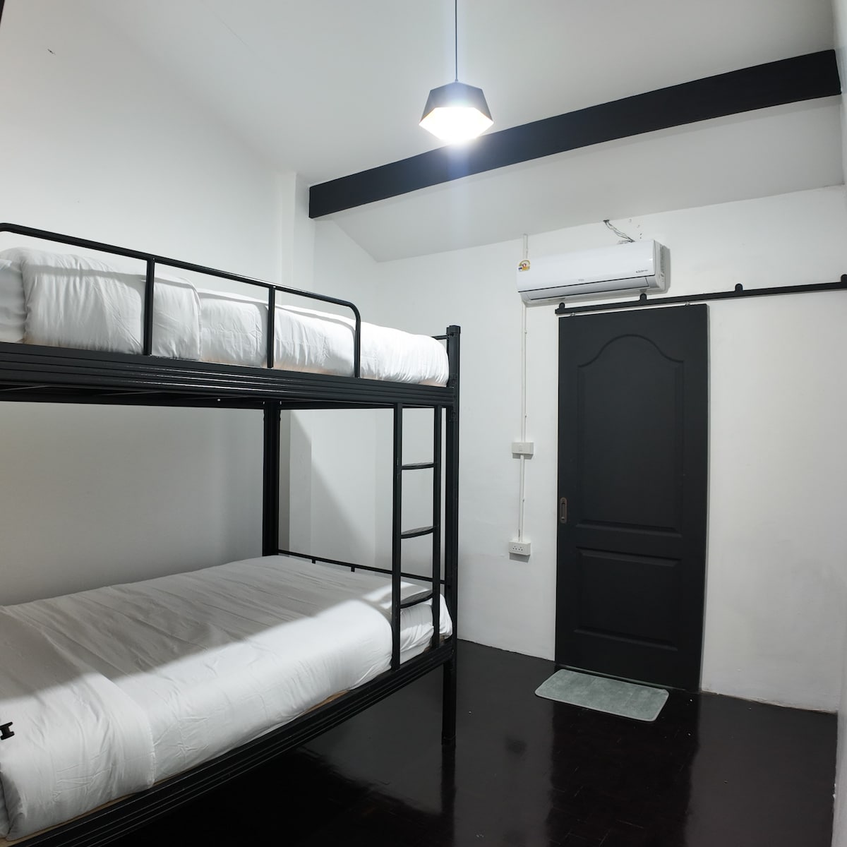 A clean and simple room features a set of black metal bunk beds with spacious mattresses. A door, painted black, provides access to the hallway. An air conditioning unit is mounted above the beds, while soft lighting illuminates the area.