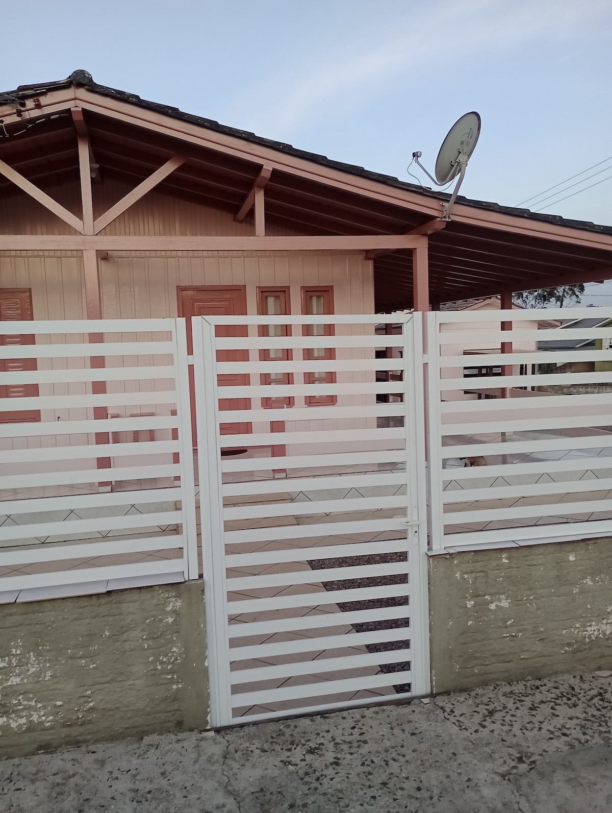 A white gate with horizontal slats is presented in front of a charming wooden house. The house features a sloped roof with dark tiles and two entry doors. A satellite dish is mounted on the roof, indicating access to media services.