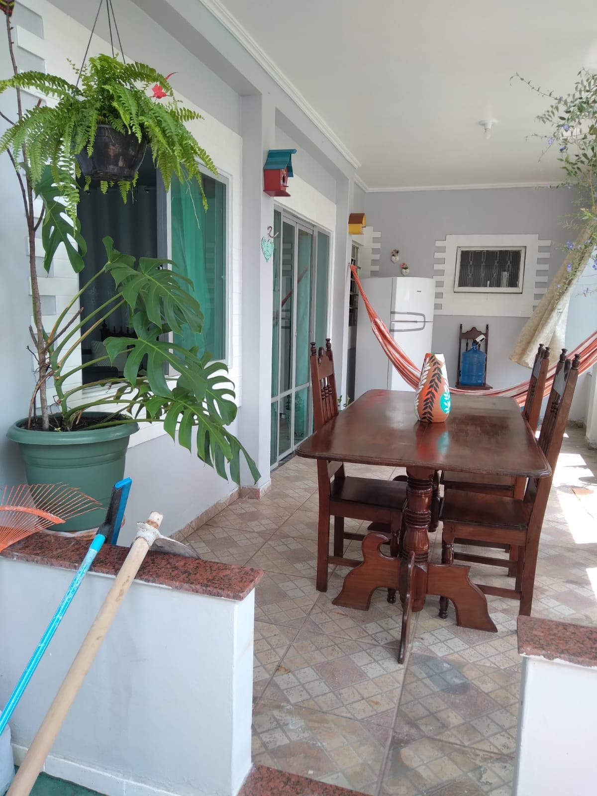 An outdoor dining area is presented with a wooden table surrounded by several chairs. A vibrant hammock is hung nearby, and decorative plants in pots add greenery to the space. A refrigerator and other furnishings are visible in the background.