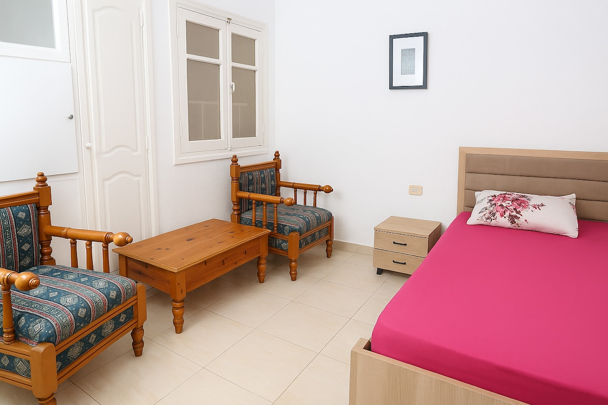 A bright bedroom features a spacious bed with a vivid pink duvet cover. Two wooden chairs with patterned upholstery and a wooden coffee table occupy the space. A small nightstand stands beside the bed, and natural light enters through a window with a simple frame.