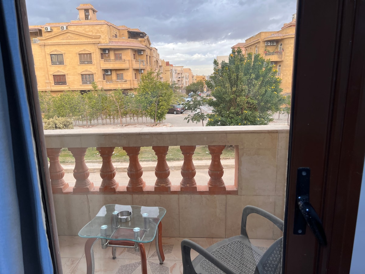 A balcony view is captured, featuring a small glass table and a chair. The surrounding neighborhood includes low-rise buildings and greenery. A pathway can be seen in front, leading to landscaped areas and additional structures.