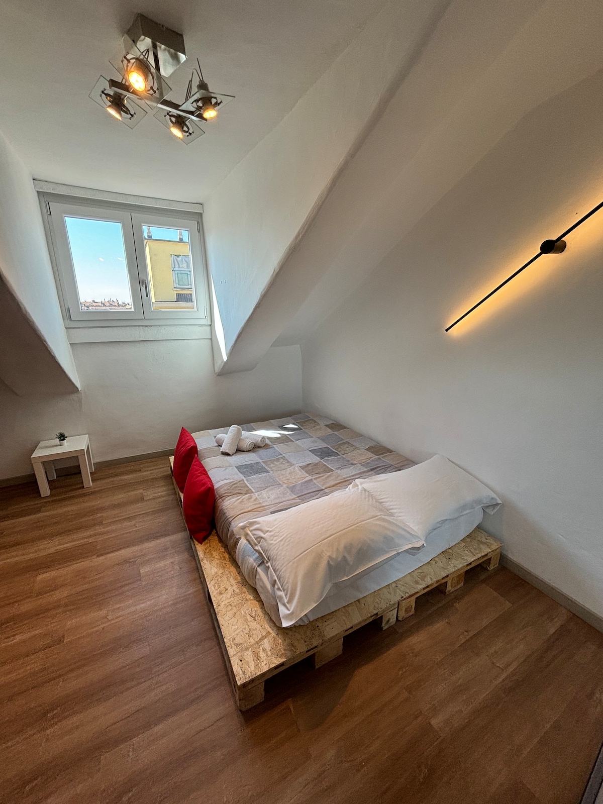 A spacious sleeping area features a comfortable bed set on a wooden pallet base, adorned with neutral-toned bedding and accent pillows in deep red. Natural light enters through a window, highlighting the room's wooden flooring and minimalist design. A small bedside table stands nearby.
