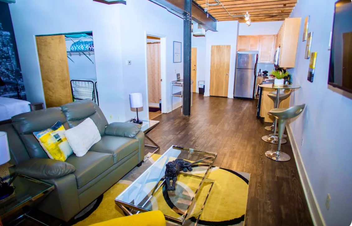 An open living area features a modern design with a comfortable gray sofa and a glass coffee table, surrounded by stylish decor. The kitchen is visible in the background, equipped with stainless steel appliances. Light wood finishes on cabinets create a warm atmosphere.