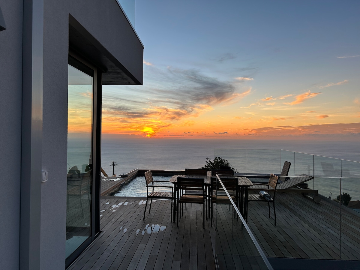 A serene outdoor space is presented with a wooden deck featuring a dining table and chairs. The setting sun casts warm hues over the ocean horizon, while a glimpse of a heated pool is visible in the background, framed by glass fencing.