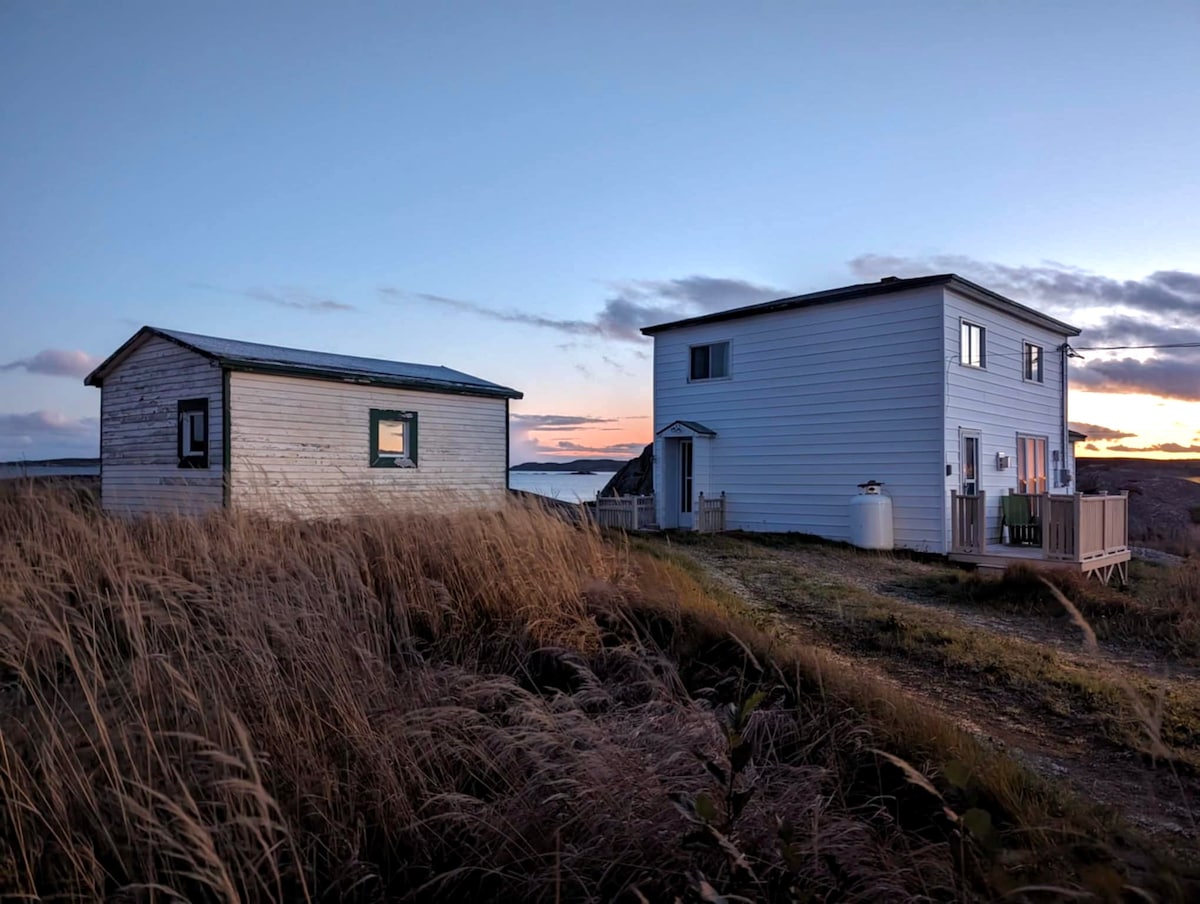 The image depicts a rustic saltbox home alongside a smaller outbuilding, both positioned on a grassy hill overlooking the sea. The sky is transitioning to dusk, showcasing soft pastel hues. The architecture reflects simplicity, emphasizing the natural surroundings.