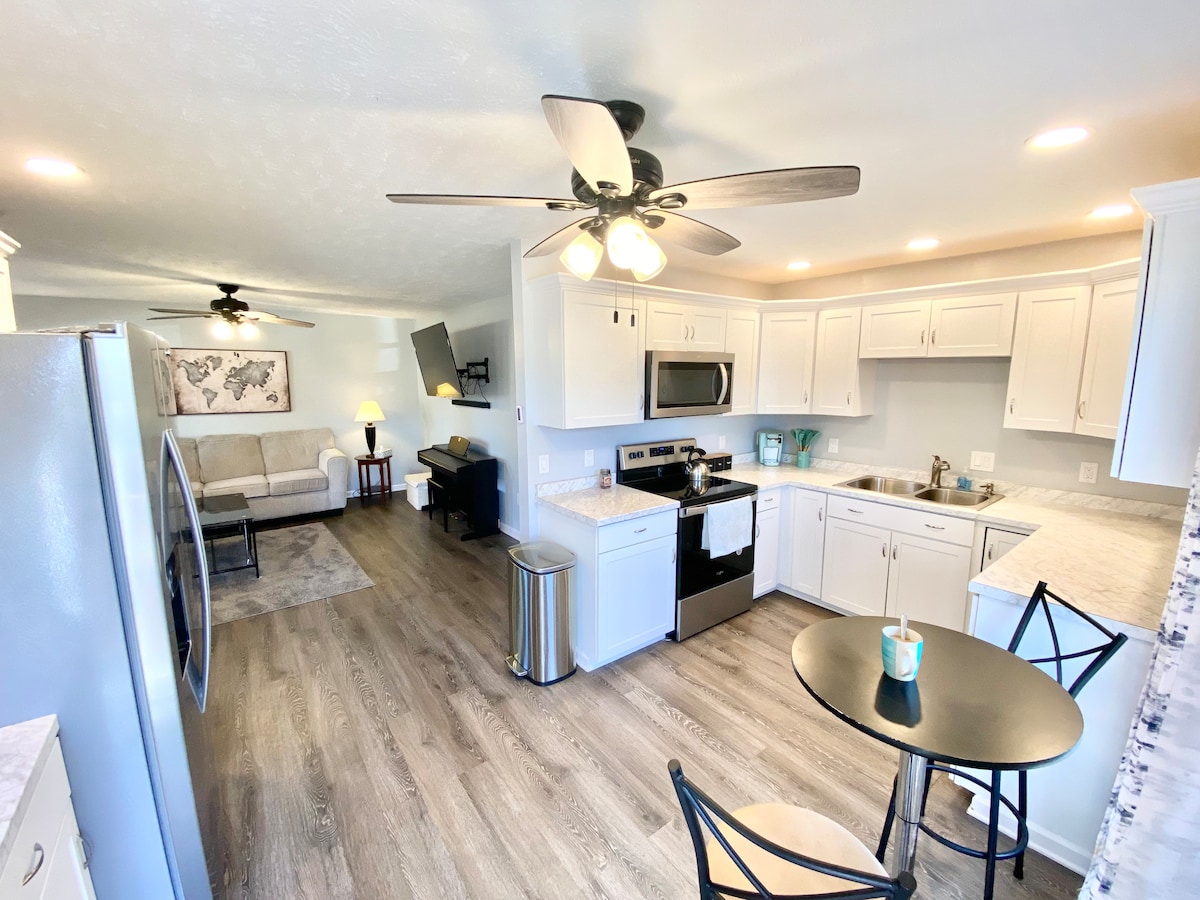 A well-equipped kitchen is displayed with modern appliances and bright cabinetry. A small round table with two chairs is positioned near the window, while the open layout reveals a cozy living area with a light-colored couch and a ceiling fan, creating a relaxed environment.