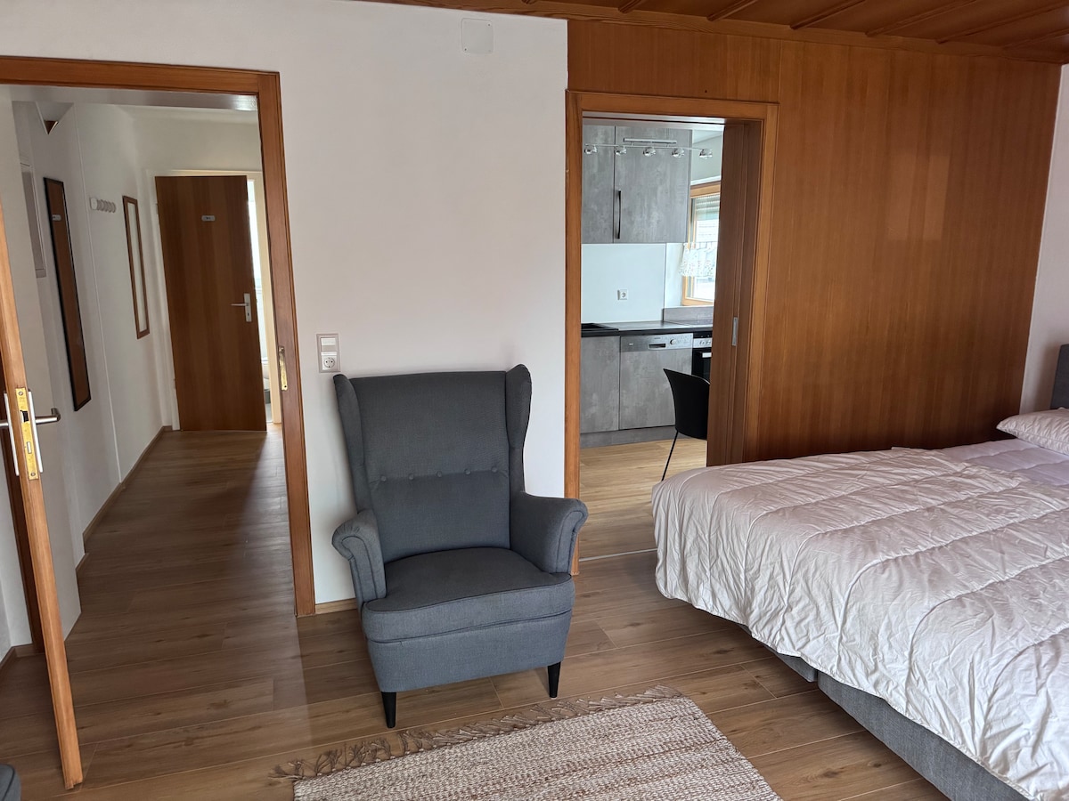 A comfortable seating area is shown with a grey armchair positioned beside a spacious bed. The warm wooden walls and flooring contribute to a cozy ambiance. A doorway leads to a kitchen area that remains partially visible, suggesting a functional living space.