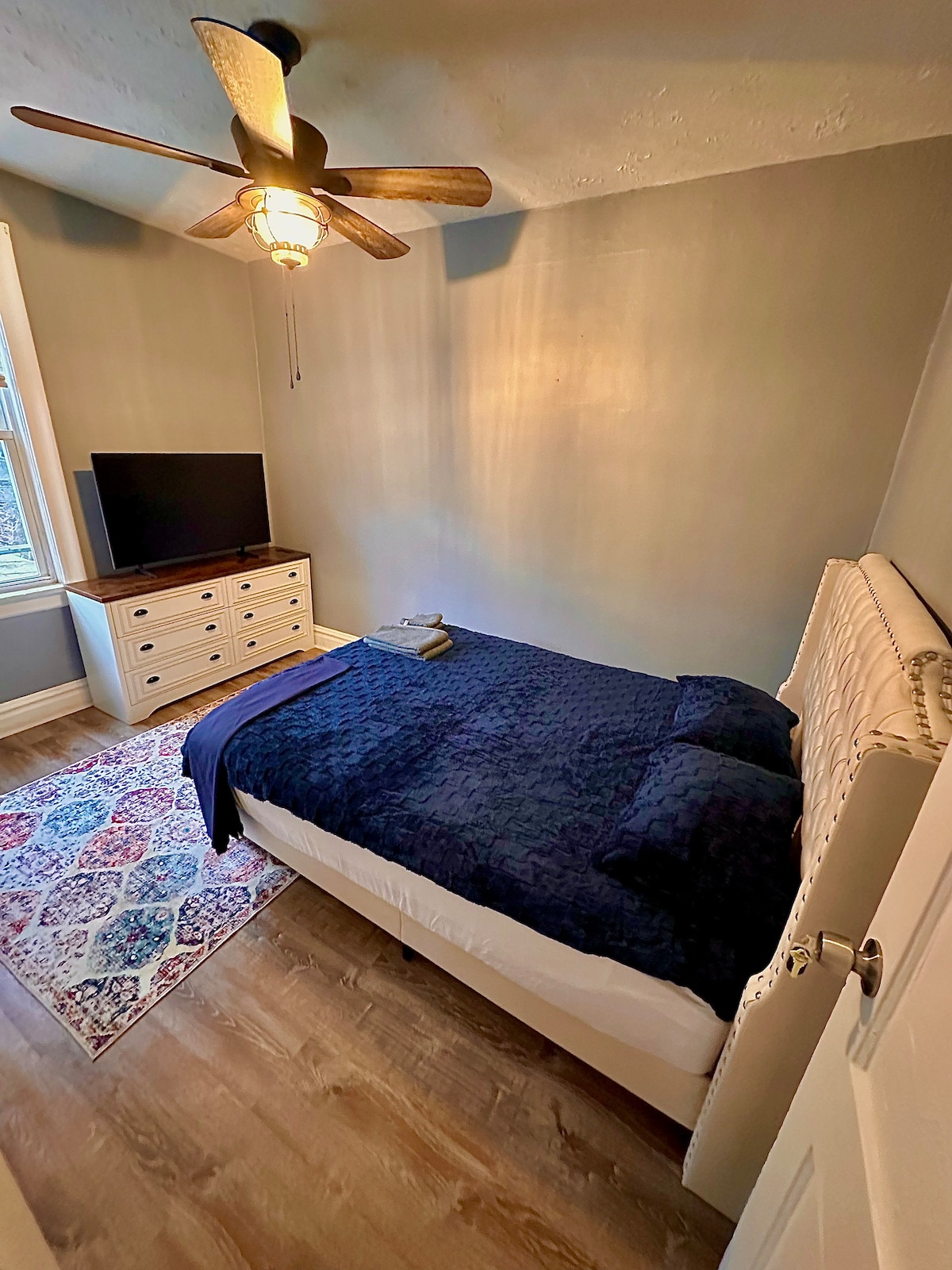 A cozy bedroom features a comfortable bed with dark bedding and two pillows. A wooden dresser and a television are positioned against the wall. A ceiling fan provides ventilation. Natural light filters through a large window, and a patterned rug covers the wooden floor.