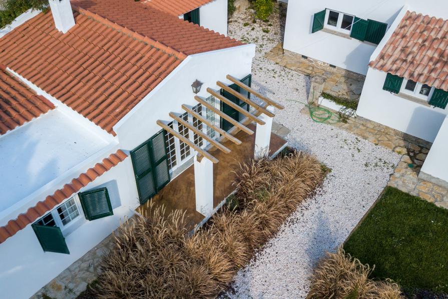 An aerial view captures the charming white house with a red-tiled roof, surrounded by well-maintained landscaping. A wooden trellis is featured at the entrance, while green shutters accentuate the windows. The gravel pathway leads towards a grassy area, enhancing the serene ambiance of the property.