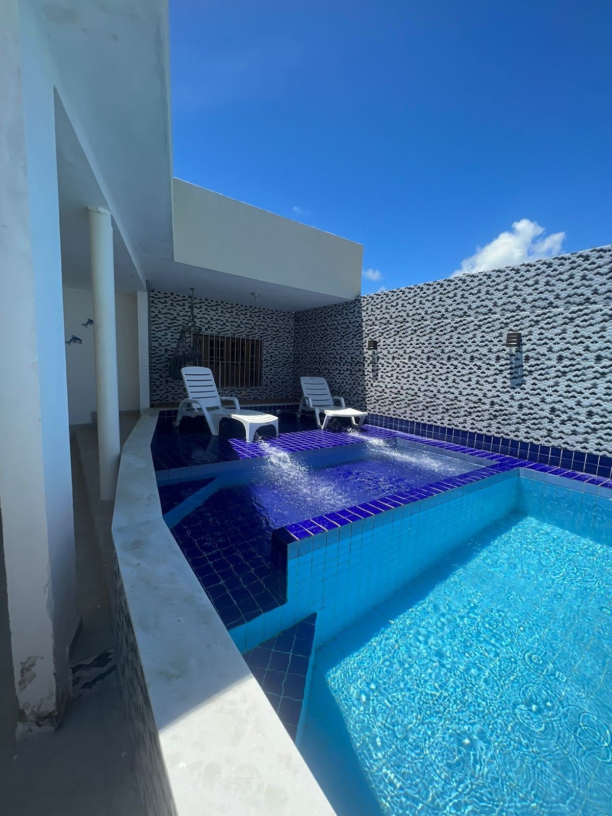 A private pool area is presented with a curved edge, featuring two white lounge chairs positioned beside the water. The pool tiles reflect a vibrant blue under the clear sky, with light streaming onto the surfaces, creating a serene ambiance.