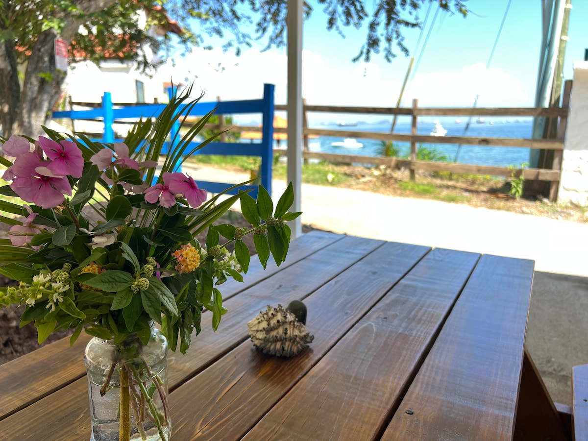 A wooden table is adorned with a simple vase holding fresh flowers, offering a touch of color. A seashell rests nearby. In the background, a view of the water and boats is visible, framed by a rustic fence and leafy trees.