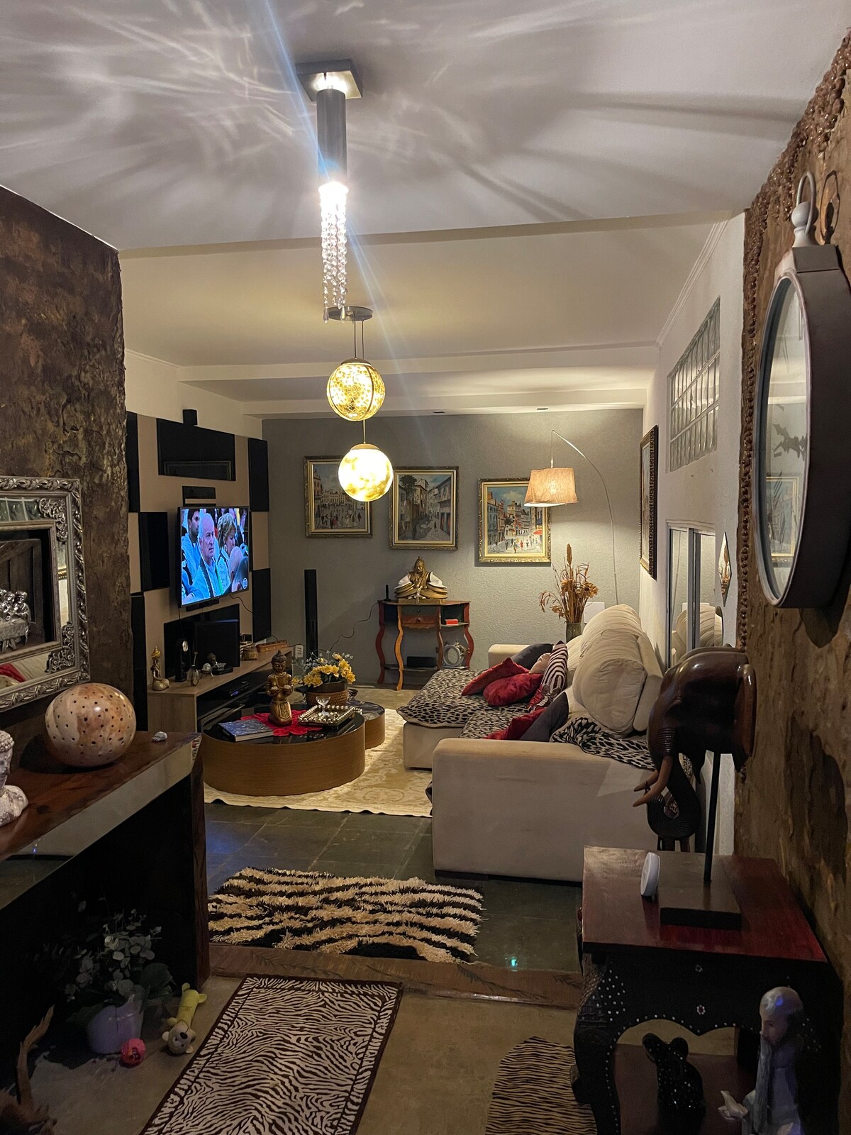 A living area features a comfortable white sofa and a coffee table, with a television displaying a vibrant image. Decorative elements like a zebra-patterned rug and wall art contribute to a cozy and unique ambiance, complemented by warm lighting from hanging fixtures.