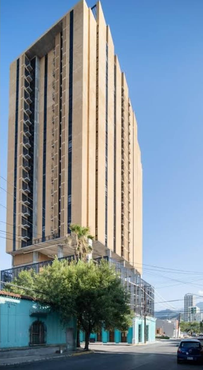 A tall, modern building stands against a clear blue sky, showcasing a sleek facade with vertical lines. The structure features numerous windows, and palm trees are visible at the base, contributing to the urban landscape.