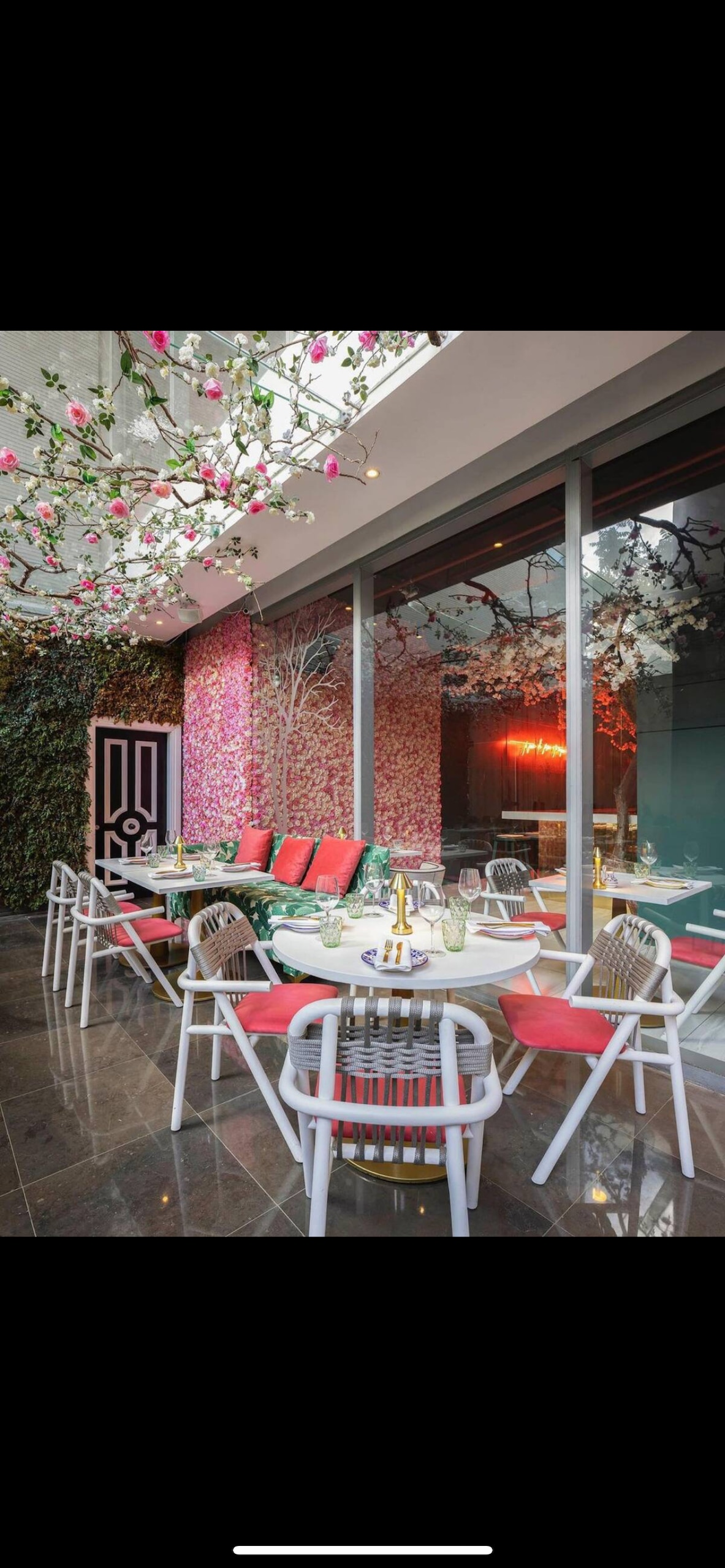 A dining area is featured with a large table surrounded by stylish chairs. The space is adorned with vibrant floral decor and greenery, creating an inviting atmosphere. Floor-to-ceiling glass walls provide abundant natural light and views of the charming outdoor space.
