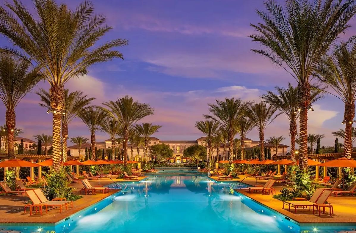 A spacious outdoor pool is framed by palm trees and lined with loungers. Orange umbrellas provide shade around the water's edge. The tranquil setting is enhanced by well-kept greenery and soft lighting, creating a serene environment for relaxation.