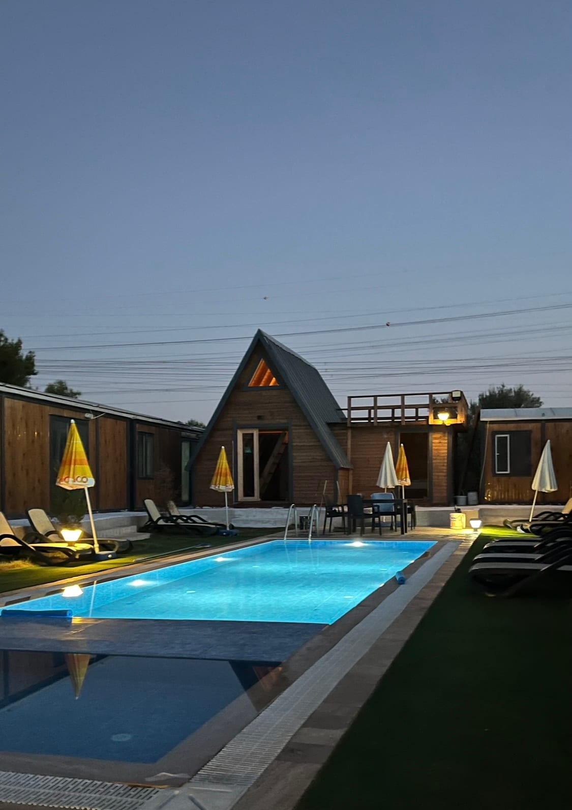 A modern pool area is surrounded by lounge chairs and umbrellas. The inviting blue water of the pool is illuminated softly at dusk, with the A-frame house visible in the background, creating a relaxing outdoor atmosphere.