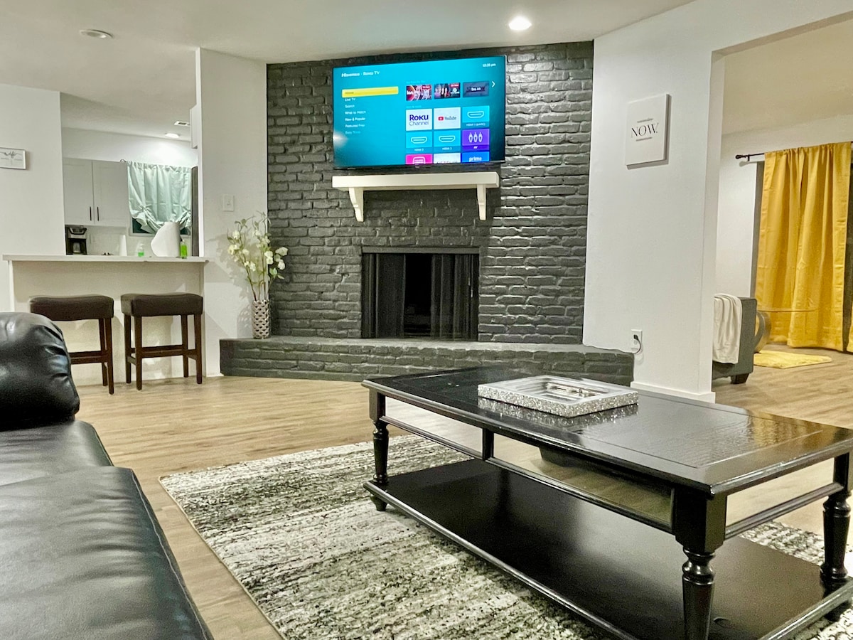 A cozy living area features a modern coffee table atop a textured rug. A sleek television is mounted above a dark brick fireplace, creating a focal point. A light-colored sofa offers comfortable seating, accompanied by two bar stools in the background near a kitchen space.