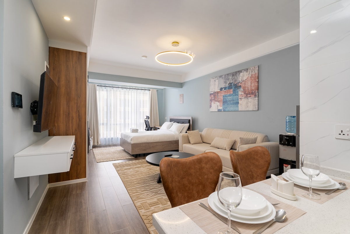 A modern studio apartment is highlighted, featuring a spacious bed and a comfortable seating area with neutral-toned furniture. A dining setup with plates and glasses is visible on the countertop. Natural light floods the space through large windows, enhancing the inviting atmosphere.
