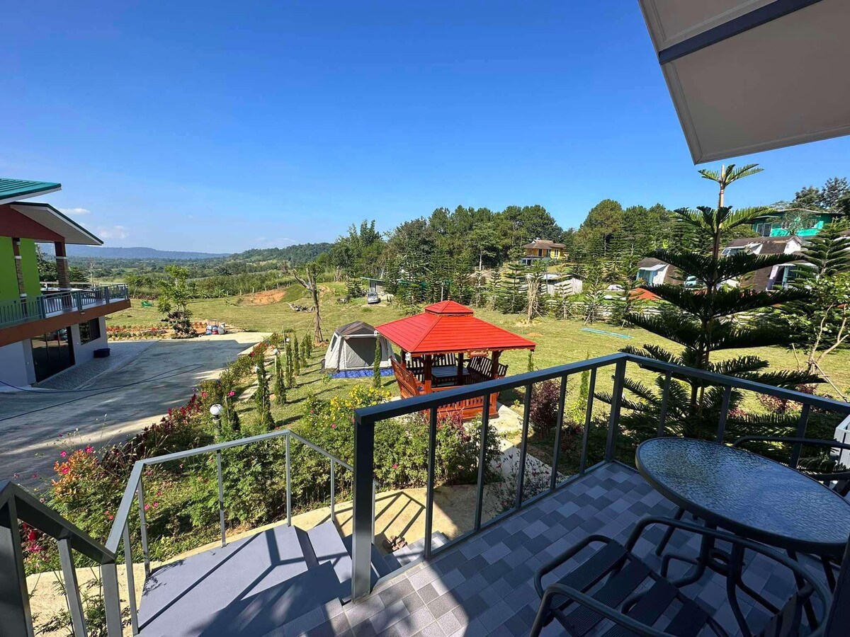A scenic view is presented from a balcony, featuring a gazebo surrounded by greenery. Tents are visible in the distance, alongside well-maintained landscaping. Sunlight illuminates the open space, creating a calm outdoor atmosphere.