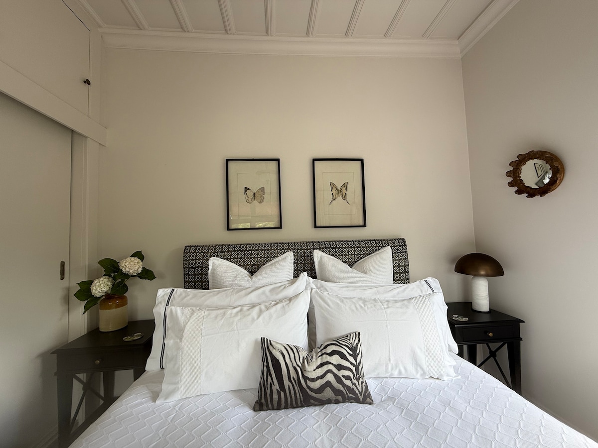 A spacious bedroom is furnished with a sizable white bed, accented by decorative pillows and a zebra-patterned cushion. Two framed butterfly artworks are displayed above the headboard. Nightstands on each side hold lamps and decorative items, while a vase with flowers adds a touch of nature.